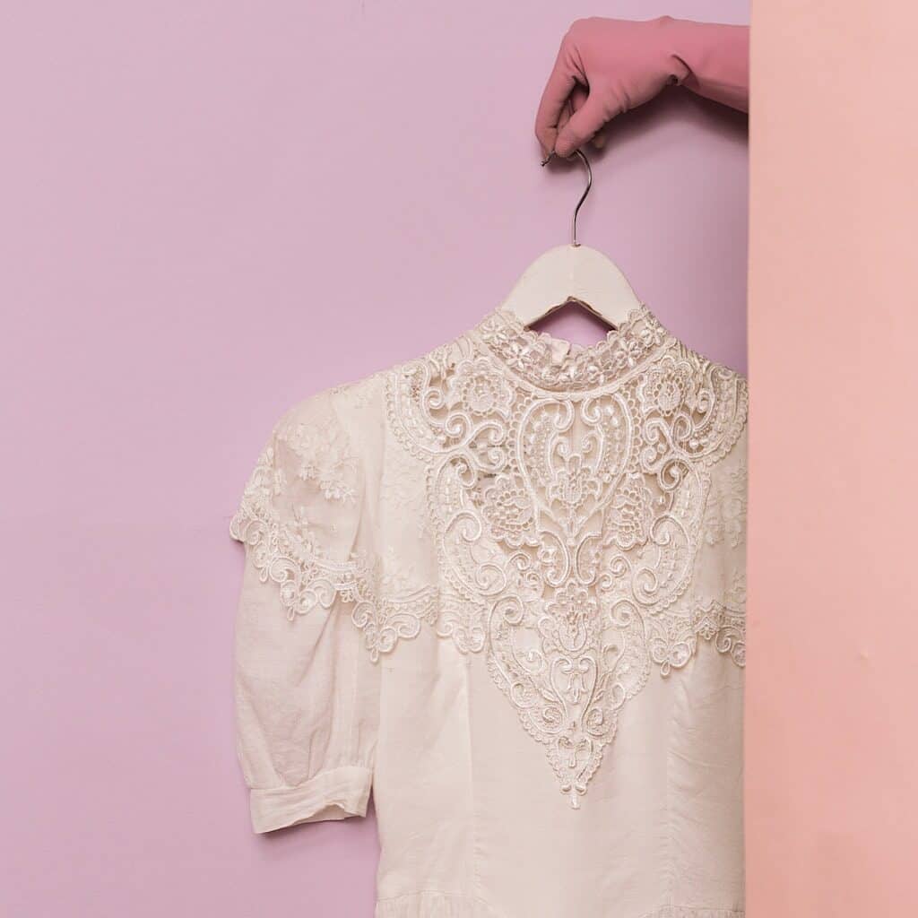 Photo shows a white blouse with lace detailing hanging on a white hanger being held by a hand wearing a pink glove against a lavender wall.