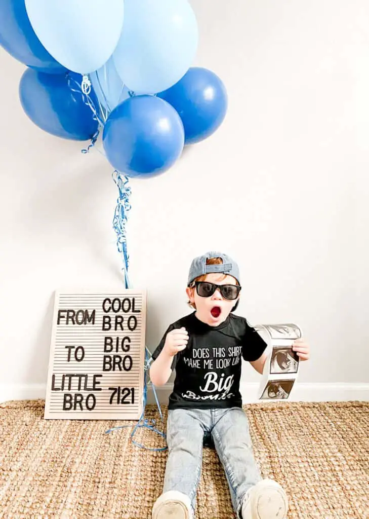 A toddler boy wearing a baseball cap backwards, black sunglasses, and a t-shirt that says "does this shirt make me look like a big brother?" He is sitting next to a letter board that says "From cool bro to big bro. Little bro 7|21>" there are blue balloons.
