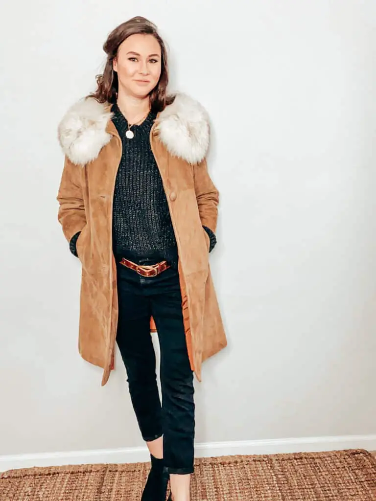 This thrifted winter outfit is made up of a camel color suede and faux fur jacket over a black knit sweater, paired with some black skinny jeans, a brown belt, a gold medallion necklace, and some black faux suede boots.