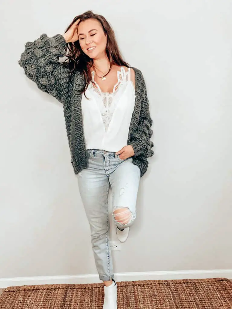 This thrifted winter outfit is a dark gray chunky knit cardigan over a white camisole with a lace panel down the front center of the top, paired with some light wash high rise jeans with some distressing and holes, a gold medallion necklace, and some white faux snakeskin ankle boots.