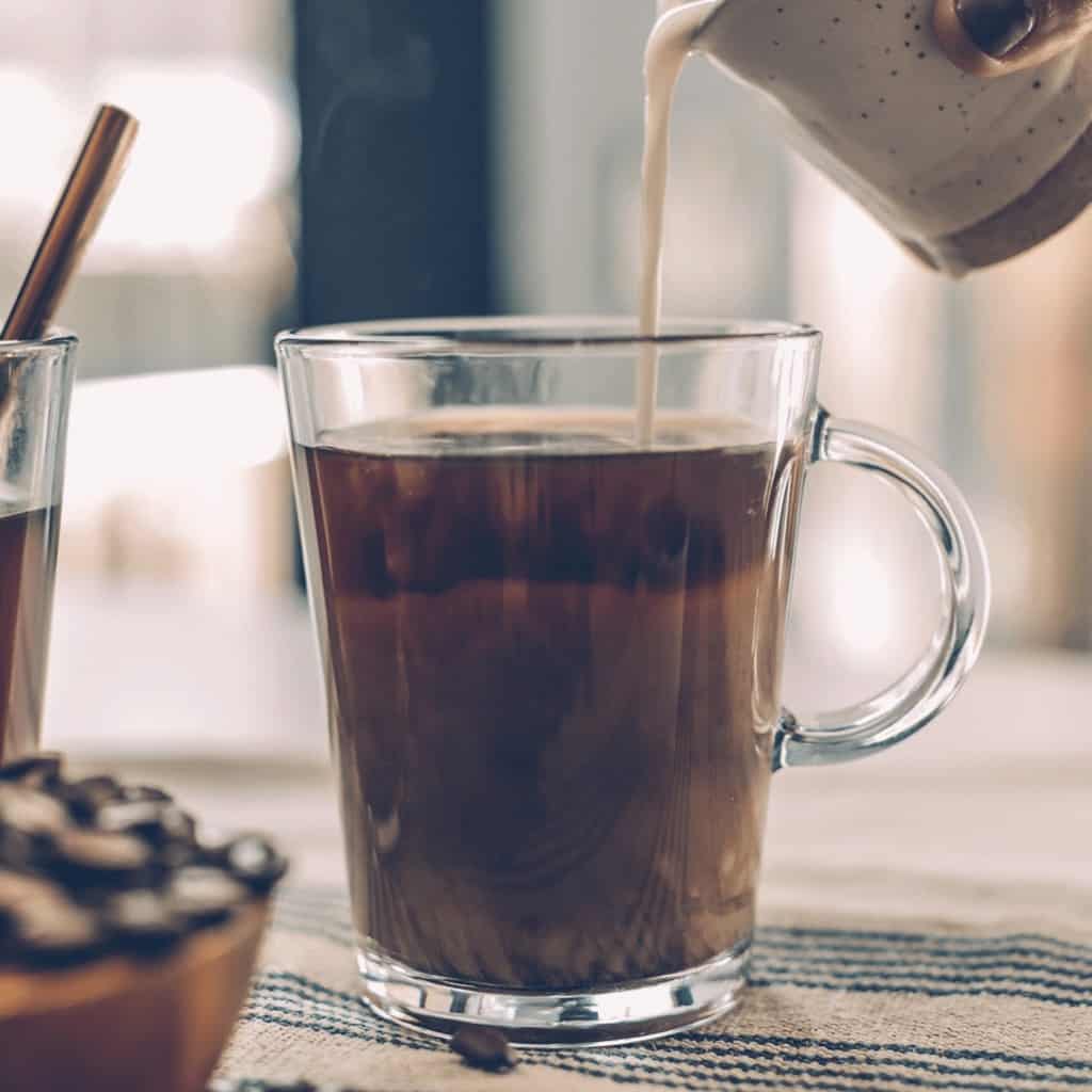 Image shows 2 clear coffee mugs filled with coffee. One mug has some creamer being poured into it. There is a bowl filled with coffee beans.