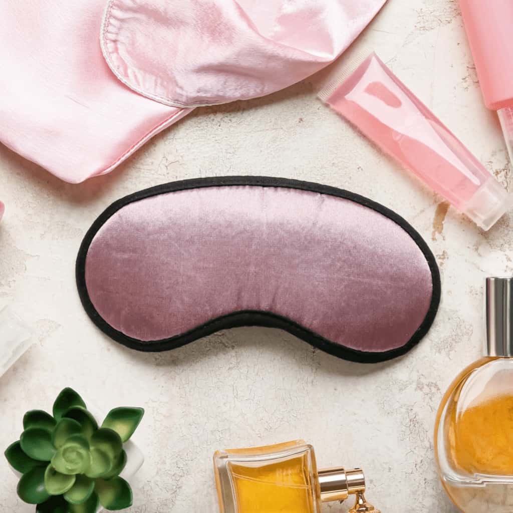 Image shows silk pajamas and an eye mask with an assortment of bottles filled with different beauty products.