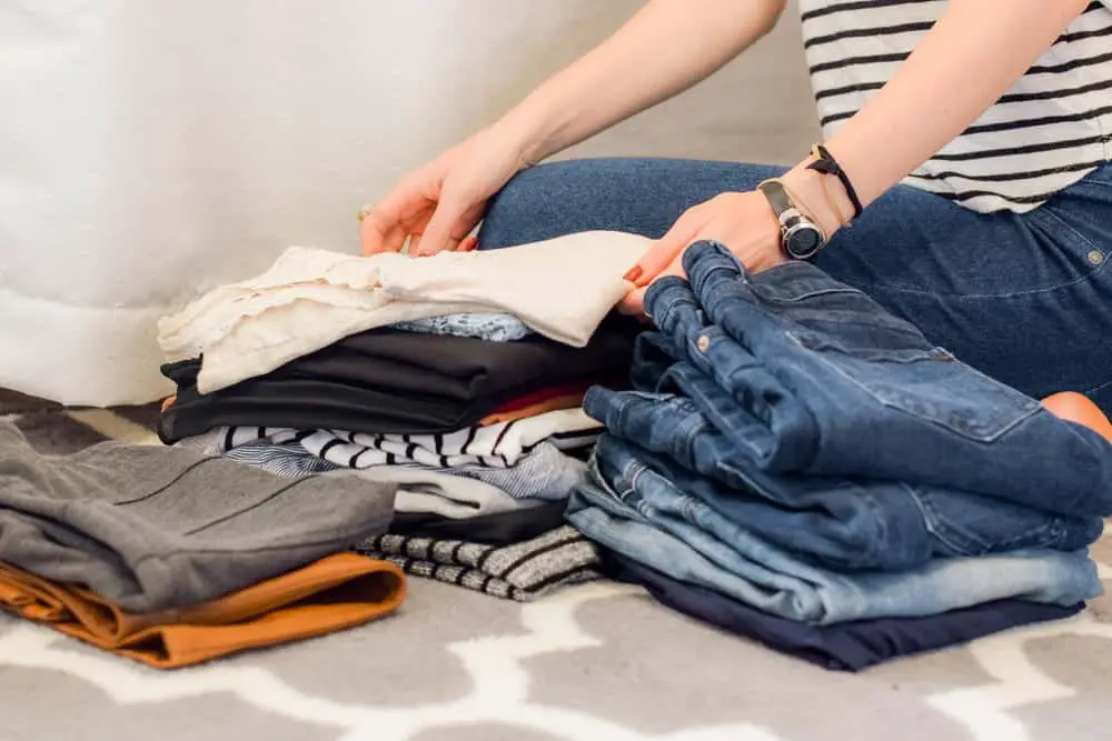 A person's torso and legs sitting on the ground sorting through small piles of folded clothing.