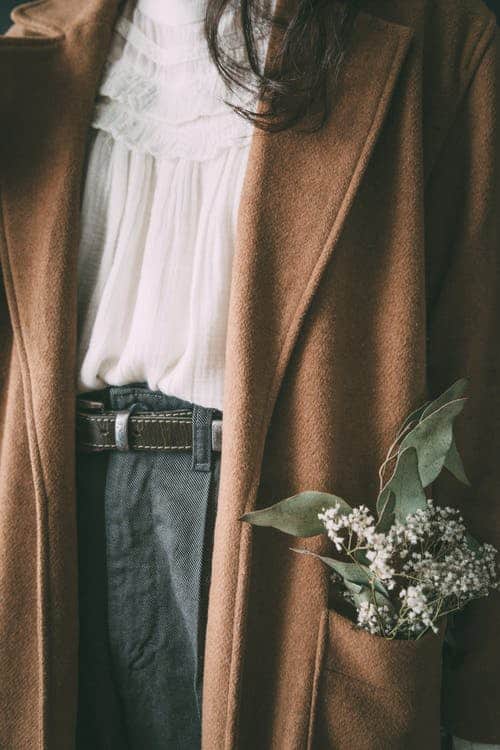 A woman's torso wearing a white top, gray trousers, and a brown wool pea coat with some baby's breath in the pocket.