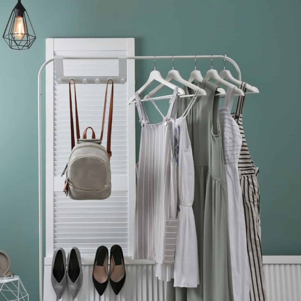 A few different style dresses in similar neutral tones hanging on a white rack along side a backpack purse and 2 sets of heels in a room with light green walls and a white door.