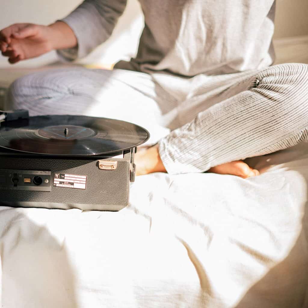 A woman sitting on a bed wearing light colored loungewear with a record player in front of her.