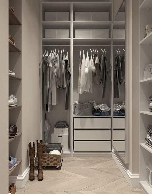 A white walk in closet with a small wardrobe in neutral tones hanging on the bar, some items are folded on shelves and there are shoes on the side wall on shelves.