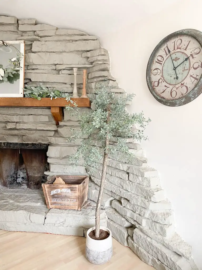 Check out how to make a fake tree for your own home that looks super realistic. Photo shows an artificial potted tree in front of a stone fireplace with a wooden box on the ledge. There is a clock on the wall, and on the fireplace mantel there is a window mirror with a spring hoop wreath on it, a garland, and 2 wooden antique looking candle holders.