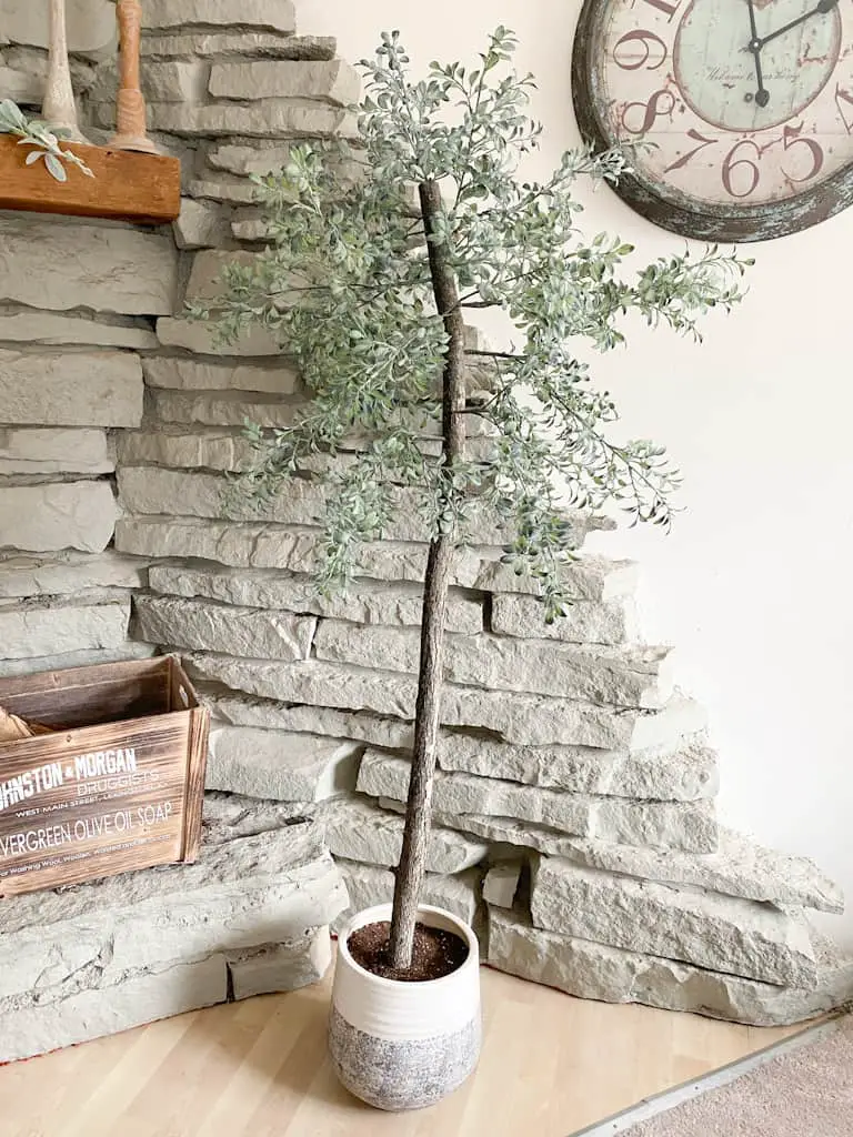 Check out how to make a fake tree for your own home that looks super realistic. Photo shows an artificial potted tree in front of a stone fireplace with a wooden box on the ledge.