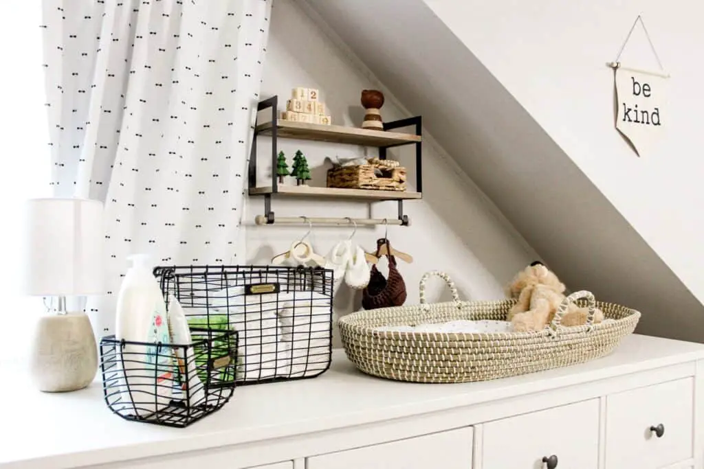 Storage is key for sharing a bedroom with baby. Photo shows a wall mounted shelf with wooden baby toys, blocks, and a small basket. Hanging on a bar under the shelf is a baby hat, knit bbay shoes, anad a wood baby teething toy all hanging on small wood hangers. On a white dresser below is a woven changing basket with a teddy bear sitting in it and some tan and a white polka dot sheet on the changing pad. Next to that are 2 different sized black wire baskets with diaper changing supplies in them.