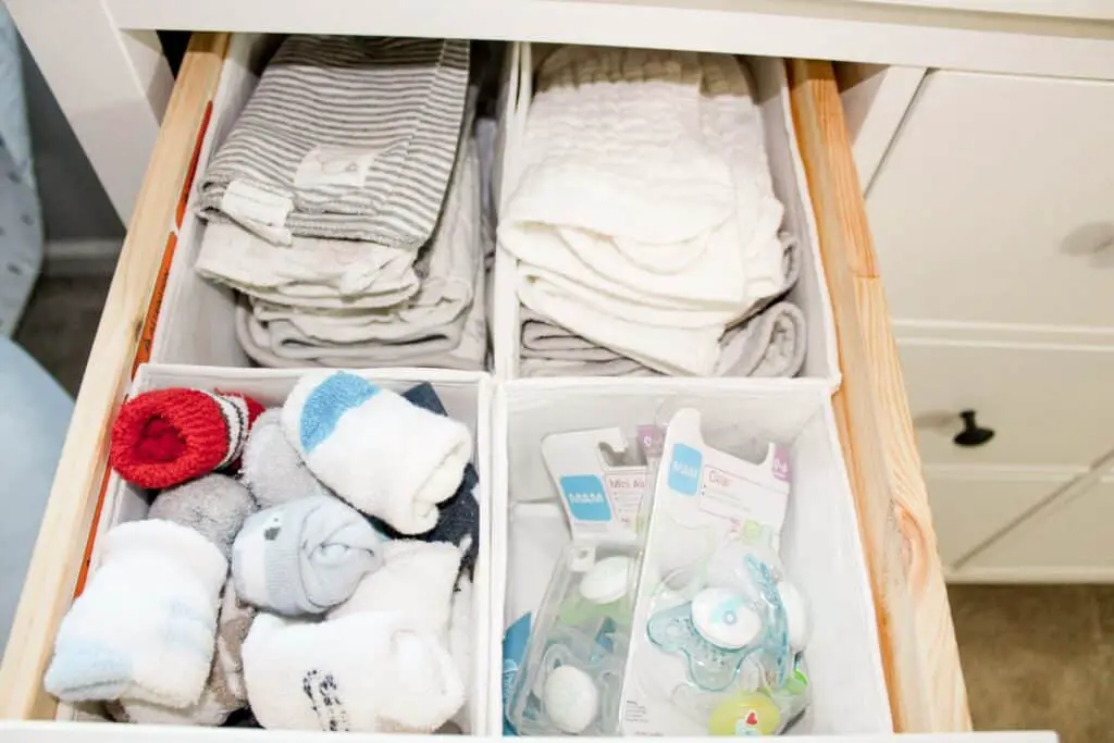 A drawer with white drawer organizers. In the organizers are burp cloths, socks, and pacifiers. Organizing is key to share a bedroom with baby.