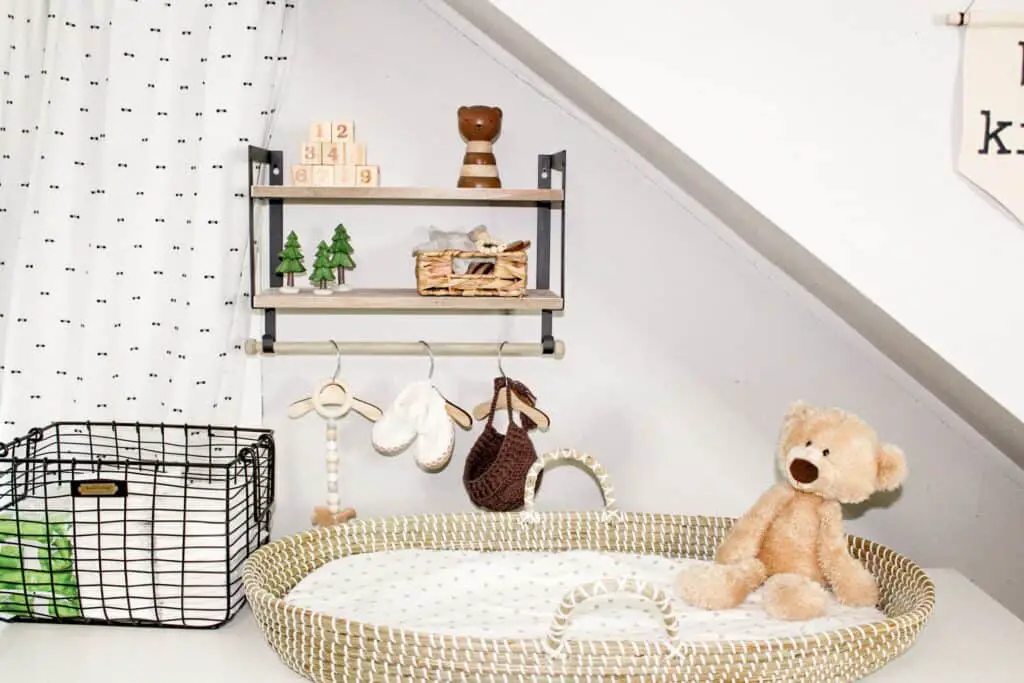 Storage is key for sharing a bedroom with baby. Photo shows a wall mounted shelf with wooden baby toys, blocks, and a small basket. Hanging on a bar under the shelf is a baby hat, knit bbay shoes, anad a wood baby teething toy all hanging on small wood hangers. On a white dresser below is a woven changing basket with a teddy bear sitting in it and some tan and a white polka dot sheet on the changing pad. Next to that are 2 different sized black wire baskets with diaper changing supplies in them.