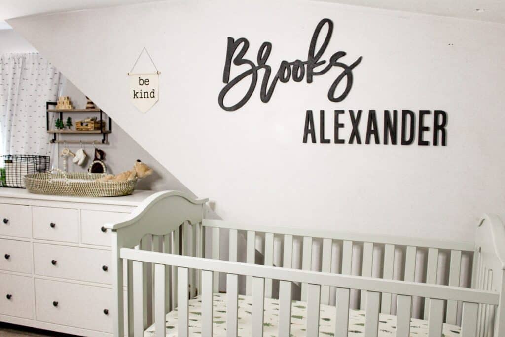 A gray crib with tree sheets, a black wood cut out of the name Brooks Alexander, a small banner that says "be kind," to the side is a white dresser, a woven changing basket, and a wood shelf with wood baby toys.