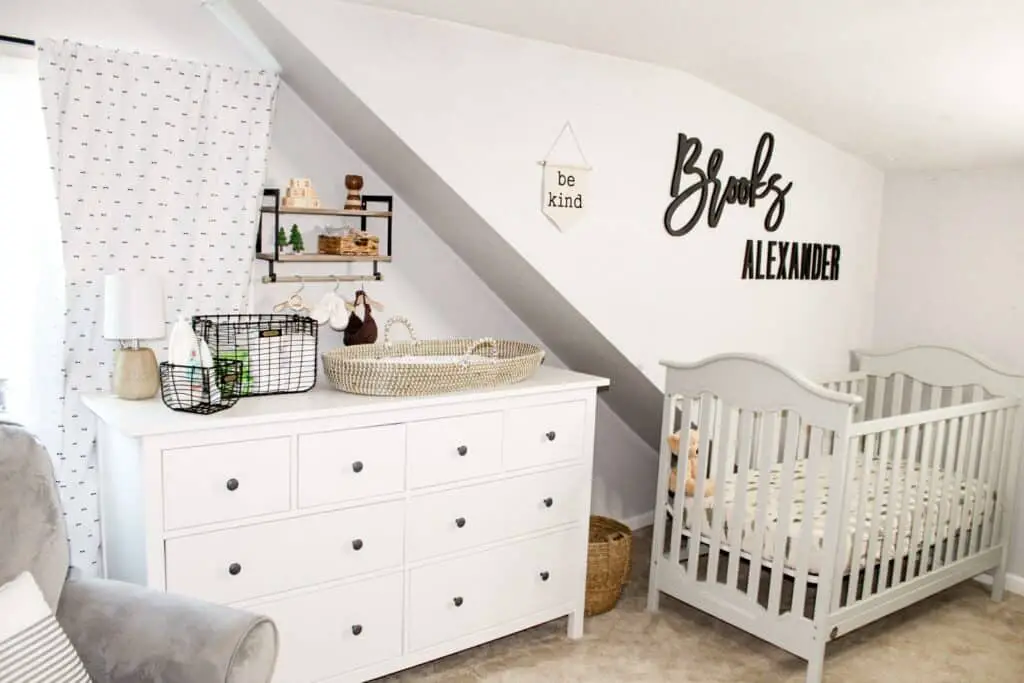 If you share a bedroom with baby then finding furniture that can house a lot of items is very helpful. Photo shows a white 8 drawer dresser with a changing station on top, a wall mounted shelf with wood baby toys and decorations and some baby accessories hanging on wood hangers on a hang bar below. There is a gray crib, a wall banner that says "be kind" and a black wood cutout of the name Brooks Alexander.