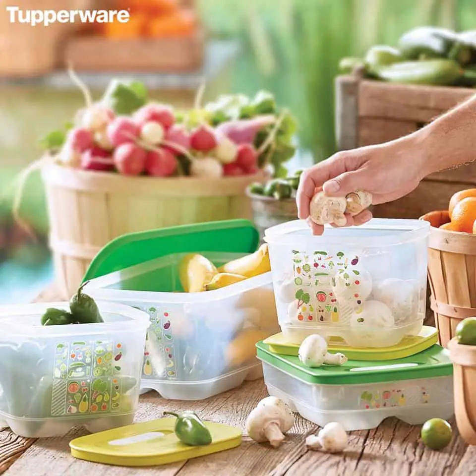 The Fridge Smart containers are also must have Tupperware products. image shows several conainers full of various produce wuth a hand reaching inside one. They are sitting on a wood table and there are wood crates of produce in the background. 