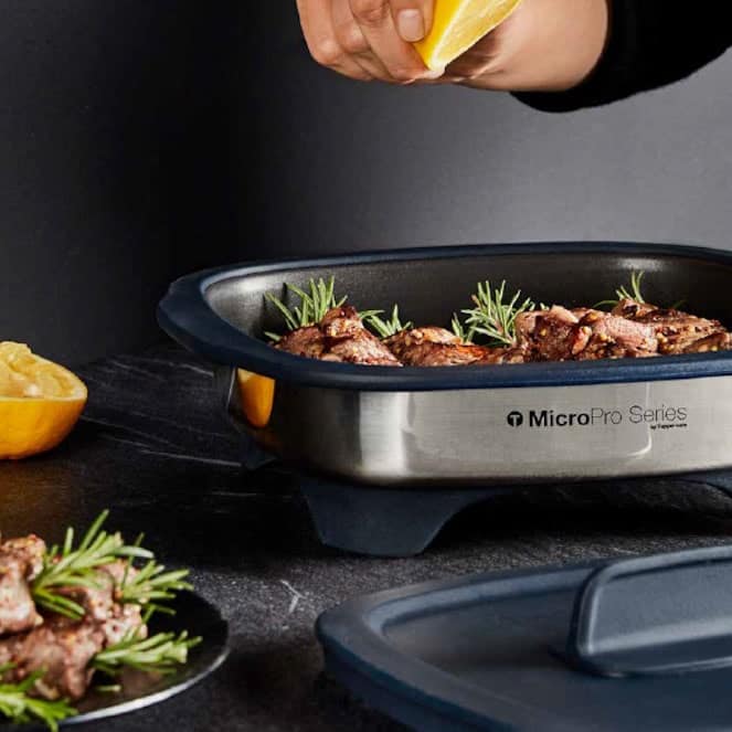 A photo of the Tupperware MicroPro Grill that has some meat and herbs inside. There is a hand squeezing a lemon over top of it. There is also a plate with the same food and the other half of a lemon sitting on a table.