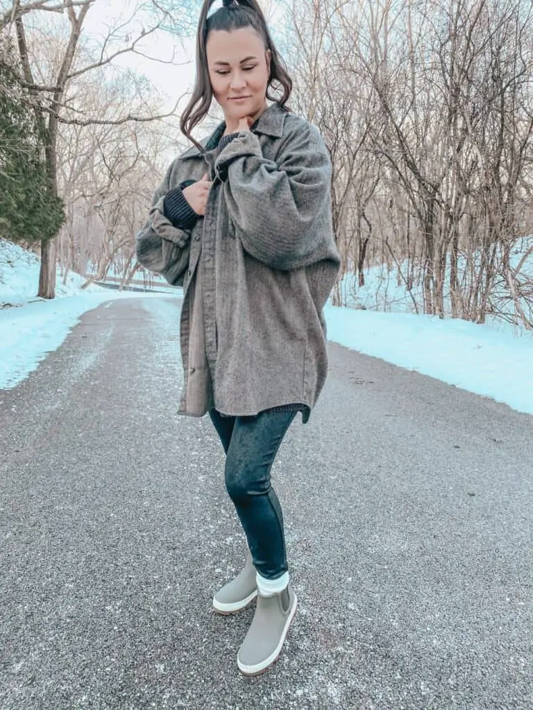 A woman posing outdoors on a street surrounded by trees. She is wearing a brown herringbone wool blend oversized button down shirt over a black long sweater paired with faux leather leggings, white socks at the ankle, and olive green Sperry rain boots.