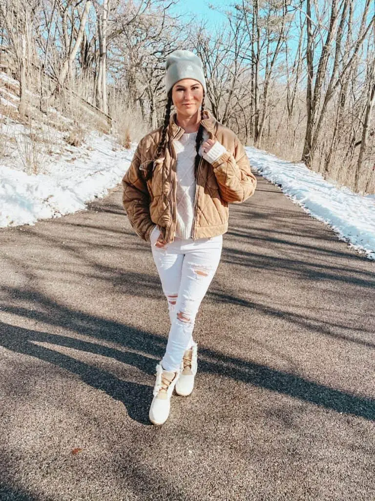 A woman posing outdoors on a street surrounded by trees. She is wearing a tan quilted jacket, a cream sweater, white distressed skinny jeans, cream and tan Sperry boots that are rubber, suede, and faux fur, and a sage green beanie.