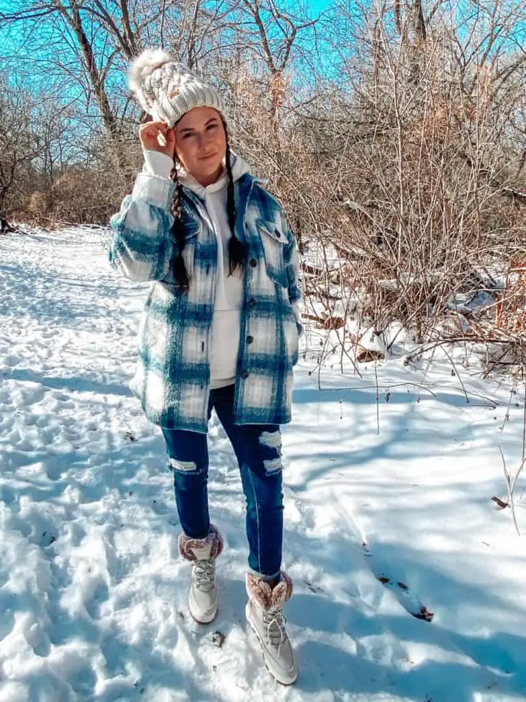 A woman posing outdoors standing in the snow surrounded by trees wearing a casual winter outfit with Sperry boots. She is wearing a blue, tan, and cream plaid shacket, a taupe hoodie, distressed blue jeans, a tan knit beanie with a cream and tan faux fur pom pom on top, and cream Sperry boots that are suede with tan faux fur on top and gold leather laces.