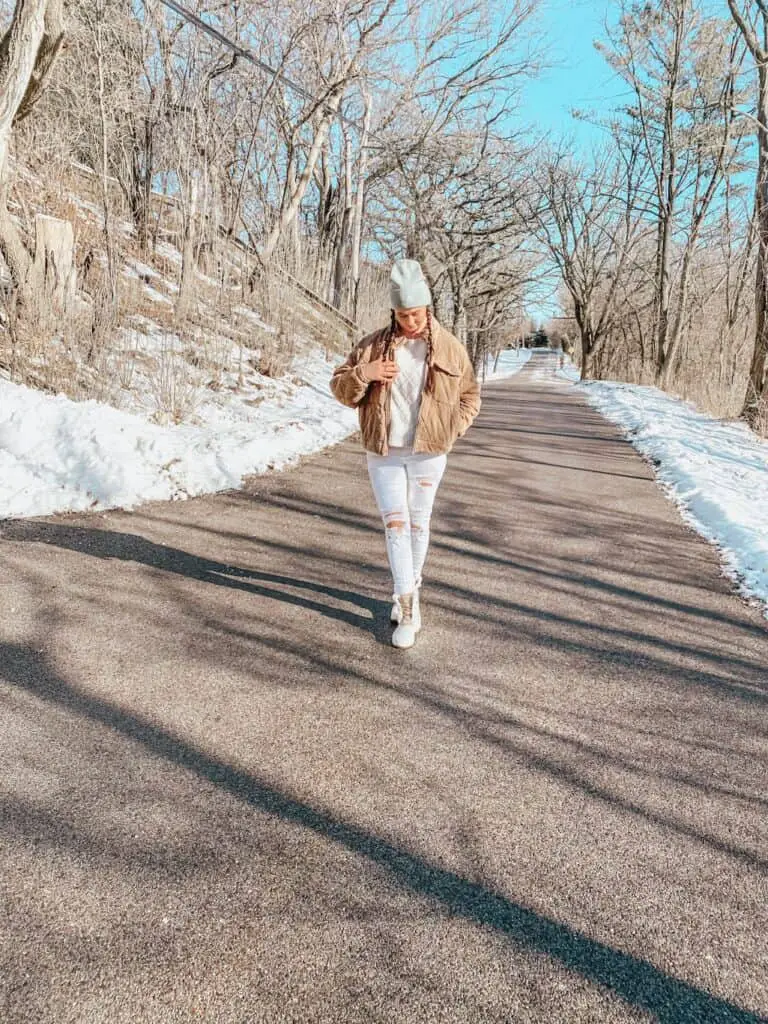 A woman posing outdoors on a street surrounded by trees wearing a casual winter outfit with Sperry boots. She is wearing a tan quilted jacket, a cream sweater, white distressed skinny jeans, cream and tan Sperry boots that are rubber, suede, and faux fur, and a sage green beanie.