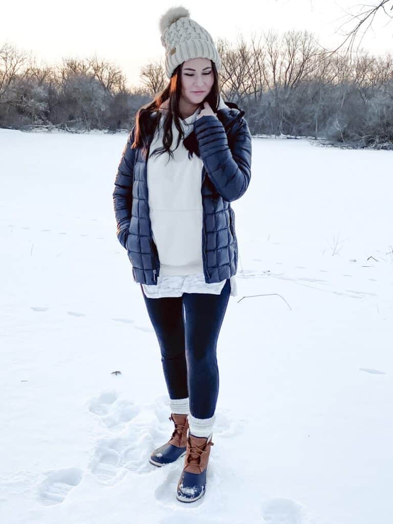 A woman posing outdoors standing in the snow with trees and a frozen pond in the background. She is wearing a black quilted puffer jacket open with a taupe hoodie layered over a long white shirt with black cotton leggings, white socks around the ankle, and a tan knit beanie with a tan and cream faux fur pom pom on top. Her boots are tan and navy leather and rubber boots with tan leather laces.