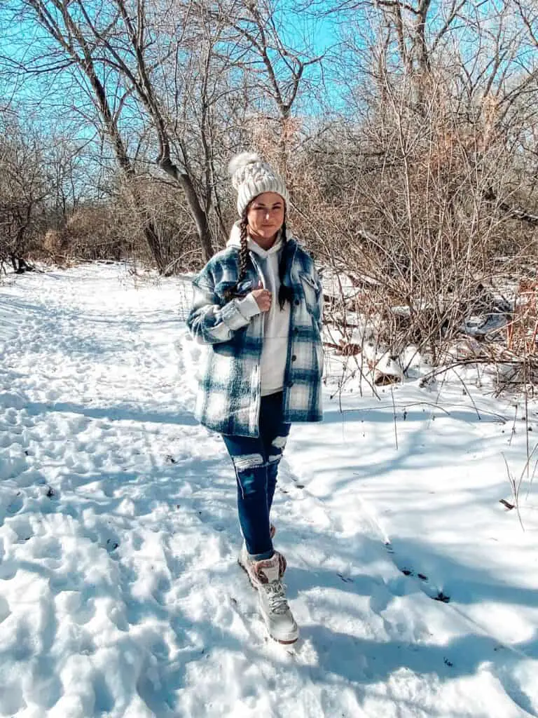 A woman posing outdoors standing in the snow surrounded by trees wearing a casual winter outfit with Sperry boots. She is wearing a blue, tan, and cream plaid shacket, a taupe hoodie, distressed blue jeans, a tan knit beanie with a cream and tan faux fur pom pom on top, and cream Sperry boots that are suede with tan faux fur on top and gold leather laces.