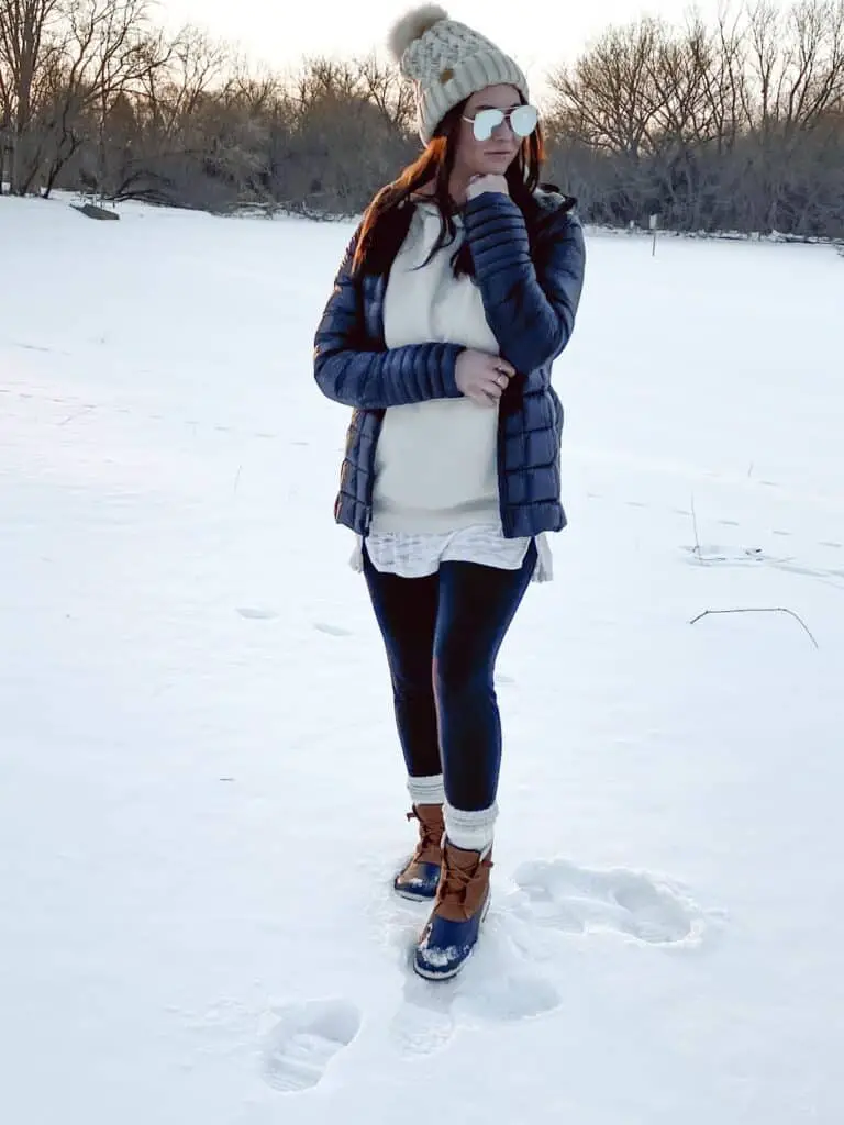 A woman posing outdoors standing in the snow with trees and a frozen pond in the background wearing a casual winter outfit with Sperry boots. She is wearing a black quilted puffer jacket open with a taupe hoodie layered over a long white shirt with black cotton leggings, white socks around the ankle, a tan knit beanie with a tan and cream faux fur pom pom on top, and some mirrored aviator sunglasses. Her boots are tan and navy leather and rubber boots with tan leather laces.