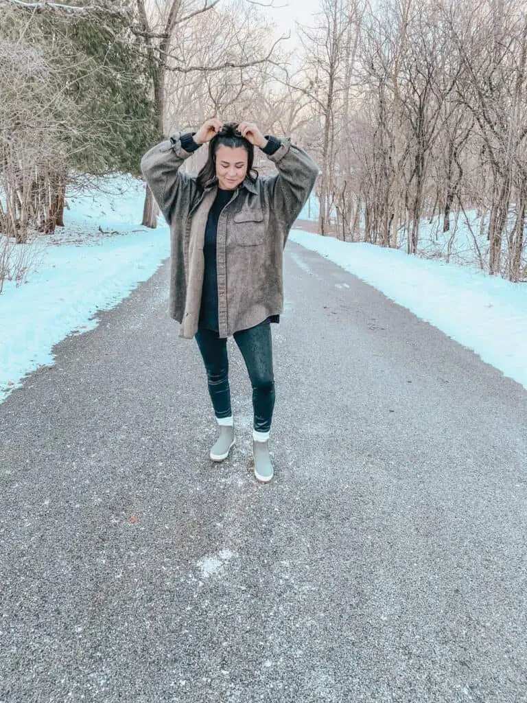 A woman posing outdoors on a street surrounded by trees wearing a casual winter outfit with Sperry boots. She is wearing a brown herringbone wool blend oversized button down shirt over a black long sweater paired with faux leather leggings, white socks at the ankle, and olive green Sperry rain boots.