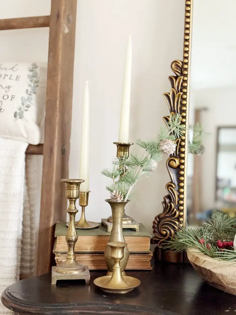 A photo of some vintage decor consisting of some assorted brass candlestick holders and 2 have white candlesticks in them, a stack of 2 old books with a faded green cover, there are some pine tree stems with small pinecones on them sticking out of a brass vase. You can see the edge of an ornate gold mirror and the edge of a worn wooden dough bowl with some pine steams with little red berries for some Christmas decor. These items are all sitting on a black tabletop.