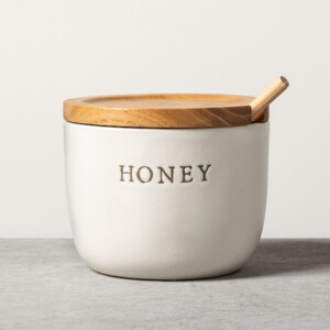 A white ceramic honey pot with the word honey engraved on it. It has a wooden lid and spoon. Perfect gift for someone who loves to decorate their kitchen.