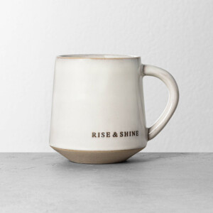 A white and tan ceramic mug with the words rise & shine engraved on it. Great to include in a gift basket for a tea or coffee lover.