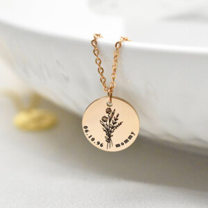 A gold round pendant necklace with flowers, a date, and the name mommy engraved on it. The necklace is hanging over the side of a white bowl. This makes a wonderful gift for her.