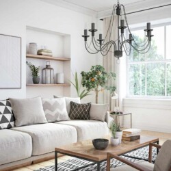 Image shows a neutral living room with a variety of decorating styles. Some modern, some boho, some farmhouse. There is a large, plush sofa with geometric patterned pillows, a metal and wood coffee table with a bowl, some potted plants, and a stack of books on it. There are some open shelves decorated with some pottery, a potted plant, a lantern, and a stack of books, There is a large black candelabra style chandelier hanging over the coffee table.