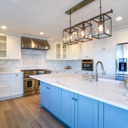 Image shows a white modern traditional kitchen. It has white cabinets with a blue island, white marble countertops, lantern style ceiling lights, marble subway tile backsplash, stainless steel appliances, and a wood floor.