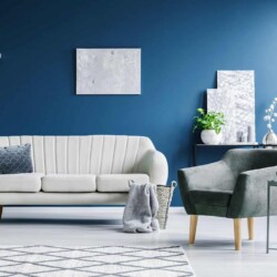 Image shows a more modern style living room with some mid century moderrn accents such as that style chair. the wall is painted a deep teal/blue color. there is a white leather sofa with a blue patterned pillow on it and in front, on the floor is a woven basket with a throw blanket hanging out of it. There is a metal table with some stacked abstract art, a potted plant, and a set of 3 different sized light gray candles on a white tray.