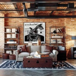 image shows an industrial style living room with a brick wall, exposed pipes and vents which are painted black, leather pillow, metal and wood accents, and a leather trunk table.