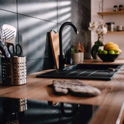 Photo shows part of a modern style kitchen with big black tiles as the backsplash, wood countertops, black metal fixtures and appliances, wood and metal accents and accessories.